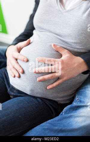 Amore, attesa, di nascita dando concetto. close up dei bracci maschio sul ventre di sua moglie incinta vestito in grigio maglione e jeans stretti, essi sono seduti che abbraccia ogni altro Foto Stock