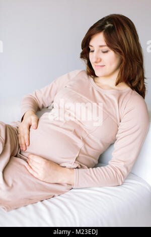 La maternità, la gravidanza, il parto concetto. sul lettino bianco è una donna straordinaria che si aspetta la madre, vestito in polvere abito ombreggiata con maniche lunghe Foto Stock