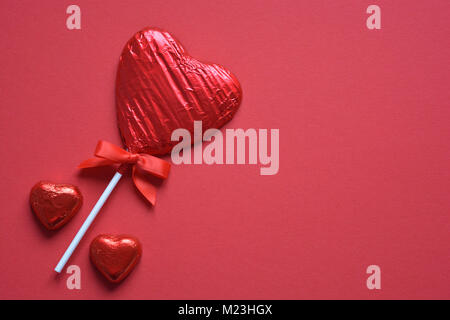 A forma di cuore, stagnola avvolto lecca-lecca di cioccolato su uno sfondo rosso Foto Stock