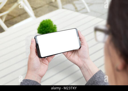 Donna che mantiene smart phone in posizione orizzontale. Schermo isolato per mockup. Bianco scrivania in legno e piante in background. Foto Stock