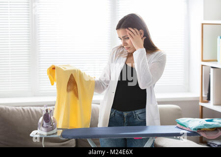 Preoccupato per il giovane donna in piedi vicino al ferro da stiro mantenendo la T-shirt con un ferro da stiro a masterizzare Foto Stock