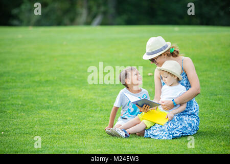 Giovane madre lettura libro ai suoi due figli piccoli seduti su erba nel parco. I bambini e la madre gode di splendida giornata estiva. Foto Stock