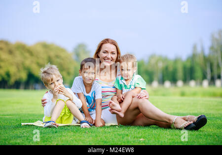 Attraente giovane donna con figli piccoli seduti su erba nel parco. La famiglia felice godendo luminosa giornata estiva. Genitori e figli relazioni. Foto Stock