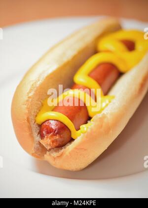 Una cotta di hot dog in un semplice panino soffice con senape su una piastra bianca. Foto Stock