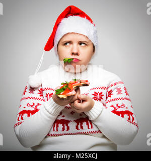 Ritratto di una donna grassa con un sandwich di grandi dimensioni nelle sue mani. Indossa una festa di Natale maglione e Santa hat. Surriscaldamenti in vacanza Foto Stock