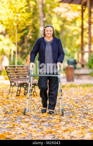 Senior donna passeggiate all'aperto con walker in autunno park e sorridente alla fotocamera. Foto Stock