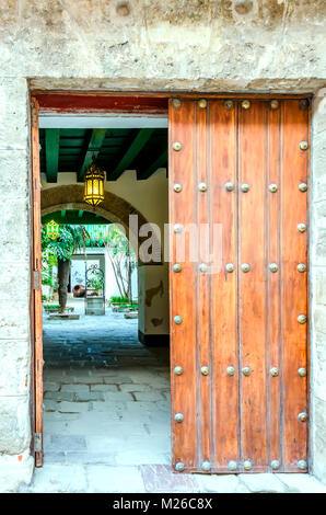 Antico cancello di legno in grigio di una parete di pietra in un cortile con un arco, luminose lampade stradali, palme, un pozzo e una brocca Foto Stock