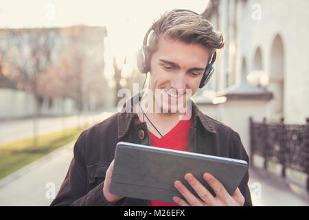 Giovane uomo informale in auricolari e refrigerazione utilizzando tablet sul background urbano. Foto Stock