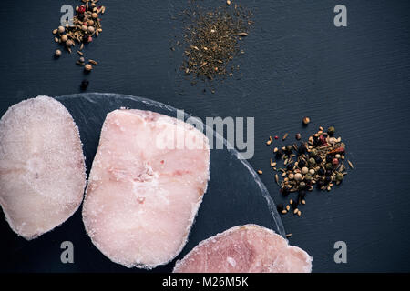 Angolo di alta vista di alcuni naselli congelati tagli su un round vassoio di ardesia, posto su un grigio scuro superficie con alcuni pali di spezie differenti, come ad esempio diversi k Foto Stock