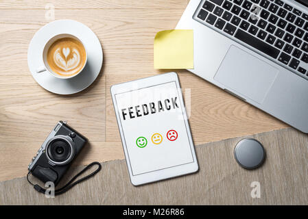 La scrittura di feedback con Emoticon colorate sulla schermata di tablet con fotocamera, tazza da caffè e laptop giacente sulla scrivania in ufficio come laici piatta Foto Stock