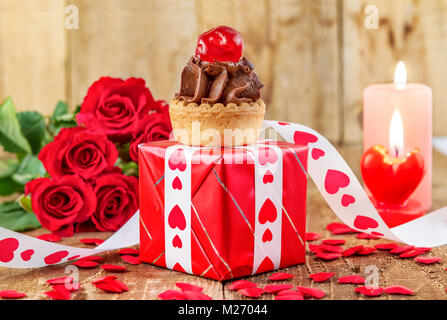 Tortina con la ciliegia sulla confezione regalo rossa nella parte anteriore del bouquet di rose rosse e candele su sfondo di legno. Il giorno di San Valentino del concetto. La festa della mamma concetto! Foto Stock