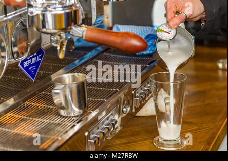 Barista versando il latte caldo in un latte di vetro con termometro latte per fare un cappuccino. Foto Stock