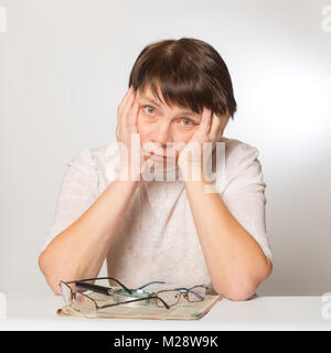 Frustrati donna mantiene la sua testa nelle sue mani. Sul tavolo di fronte a lei è un giornale, due bicchieri e una lente di ingrandimento. Scarsa visione, occhiali ar Foto Stock