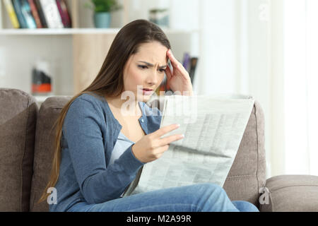 Ritratto di una donna in questione la lettura di cattive notizie in un giornale seduto su un divano nel salotto di casa Foto Stock