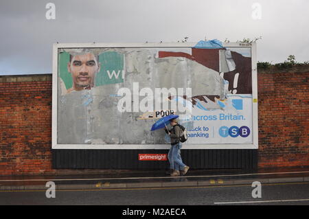 Due persone che camminano con gli ombrelli sotto la pioggia davanti ad un cartellone pubblicitario in Bristol, Regno Unito Foto Stock