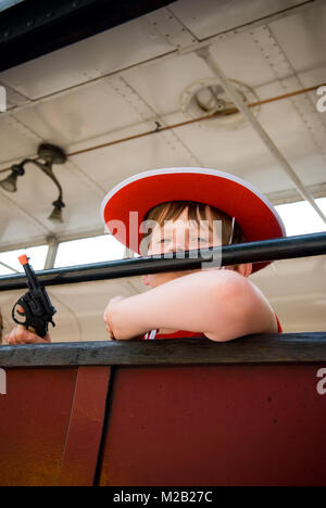 Cerca fino a 8 anno vecchio ragazzo vestito da cowboy e azienda pistola giocattolo appoggiata al di fuori del vecchio treno finestra Foto Stock