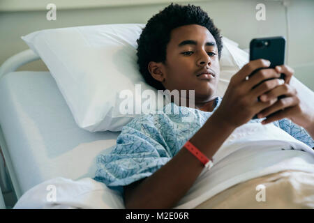 Black Boy che stabilisce nel letto di ospedale sms sul cellulare Foto Stock