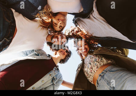 Ritratto di sorridere giovani amici formando un huddle contro sky. Allegro gruppo di uomini e donne amici huddling all'esterno. Foto Stock