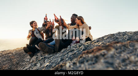 Variegato gruppo di amici seduti sulla cima della montagna e birre di tostatura. Allegro giovani uomini e donne godendo con birre all'aperto al tramonto. Foto Stock