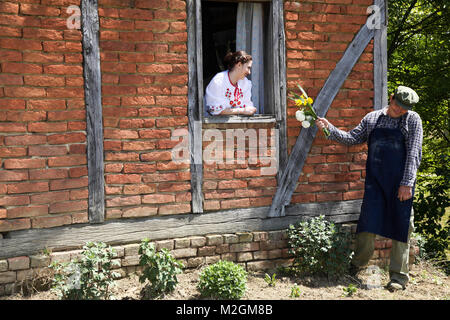 La ragazza sulla finestra di una vecchia casa di mattoni, tradizionalmente condita, il ragazzo timidamente dà il suo bouquet di fiori Foto Stock