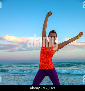 Aggiornamento wild lato mare allenamento. A piena lunghezza Ritratto di felice donna attiva nell'abbigliamento sportivo in riva al mare al tramonto gioia Foto Stock