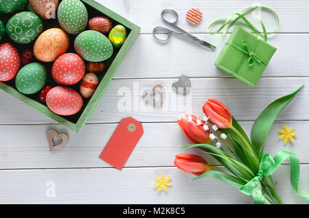 Vista dall'alto su bianco tavolo in legno con scatola del dipinto di uova di Pasqua, arancio tulipani e confezione regalo. Artigianato pasqua concetto. Foto Stock