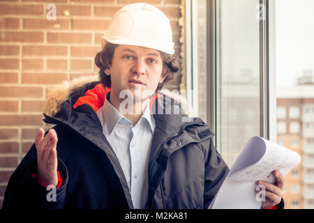 Un arrabbiato scontenti builder lavoratore in un casco con disegni di progetto prevede nel suo una mano e telefono cellulare in un'altra mano urla a qualcuno. Bad e Foto Stock