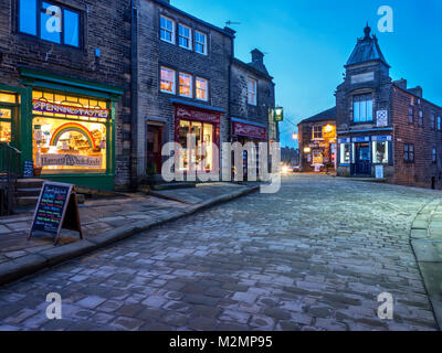 Le luci provenienti lungo la strada principale lastricata in pietra in Haworth Village al crepuscolo West Yorkshire Inghilterra Foto Stock