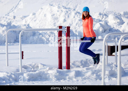 Giovane donna in un blu brillante cappello, maglione arancione e elk rende pull-ups sulle barre orizzontali su un luminoso giorno di inverno Foto Stock