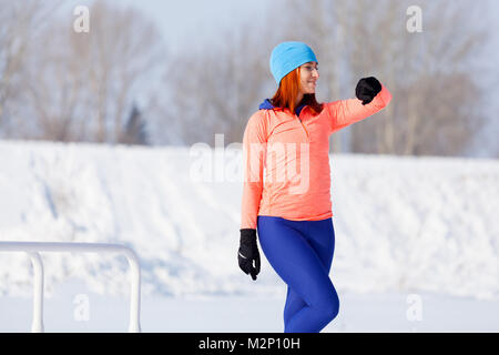 Una giovane donna in un blu brillante cappello, maglione arancione e elk sorrisi, si prepara per la formazione e la guarda l'orologio sportivo prima di eseguire sul playgroun Foto Stock