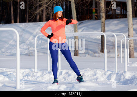 Una giovane donna in un blu brillante cappello, maglione arancione e elk sorrisi, si prepara per la formazione e la guarda l'orologio sportivo prima di eseguire sul playgroun Foto Stock