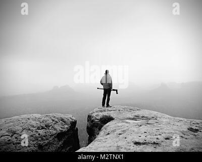 Escursionista con la fotocamera sul cavalletto prende foto dalla vetta rocciosa. Fotografo solo in corrispondenza del bordo fotografia brumoso paesaggio, foresta in valle. Foto Stock