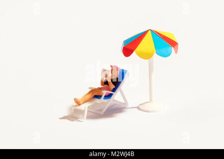 Persona in miniatura per leggere un libro in relax sulla sedia reclinabile a prendere il sole in ombra sotto un colorato ombrellone su bianco con spazio di copia Foto Stock