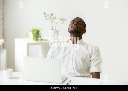 African lavoratore di ufficio rilassante con gli occhi chiusi seduto alla scrivania con computer portatile, nero dipendente tenendo il resto facendo esercizi per il rilassamento a workplac Foto Stock