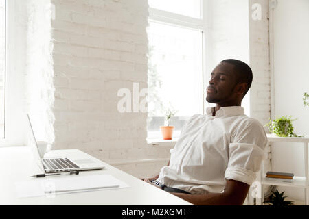 African American uomo rilassarsi dopo il lavoro respirando aria fresca seduti a casa scrivania da ufficio con computer portatile, nero rilassato imprenditore meditando con gli occhi Foto Stock