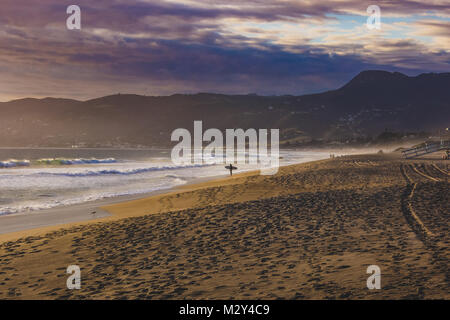 Surfista maschio permanente sulla spiaggia tenendo la sua tavola da surf a Point Dume State Beach con le nuvole colorate nel cielo e Santa Monica Montagne in ba Foto Stock