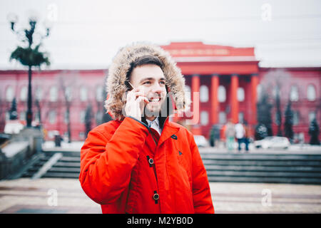 Ragazzo con la barba e la giacca rossa nel cofano uno studente utilizza il telefono cellulare e tiene in mano vicino alla testa, parlando al telefono con il sorriso sulla backg Foto Stock