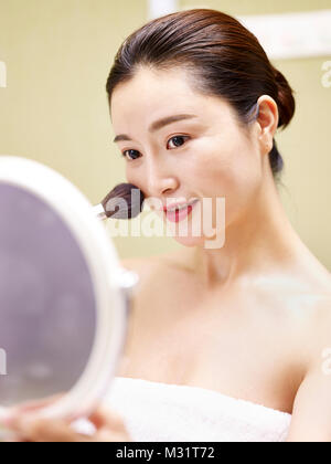 Bella giovane donna asiatica guardando nello specchio mentre si applica il make-up sul viso utilizzando una spazzola. Foto Stock