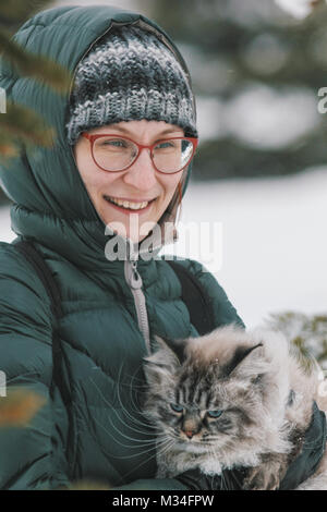 Giovane donna in verde down jacket e bicchieri tenendo un grande cat nella foresta di inverno Foto Stock