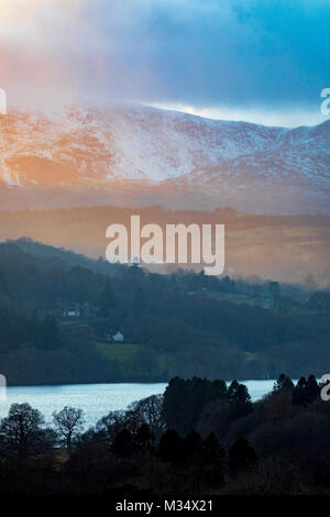 Lago di Windermere, Cumbria. Regno Unito Meteo. Bella fine ad una bella giornata nel distretto del lago come il sole tramonta oltre le montagne e il lago Windermere con neve ancora presente sulle cime Foto Stock