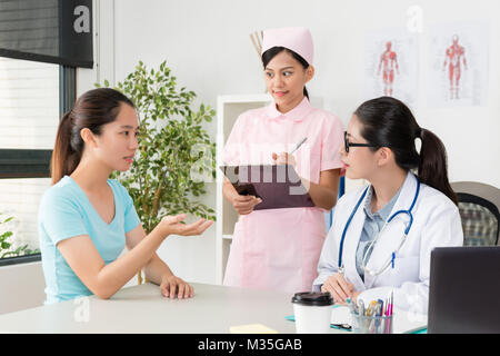 Giovane e bella infermiera in piedi in ufficio la scrittura di record medico sul noteboard quando il paziente che illustra personal malati problema per il medico. Foto Stock