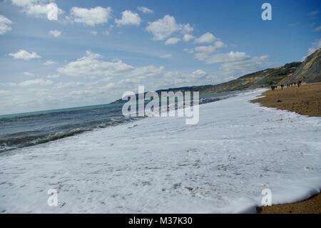 Swasing calma onda sulla spiaggia Charmouth, guardando ad ovest verso Lyme Regis città sotto un Cumulus blu cielo velato. Il West Dorset, Regno Unito. Foto Stock