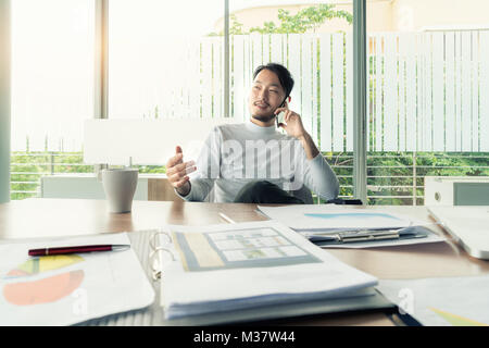 Ritratto di asiatici imprenditore sorridente o architetto parlando sul telefono cellulare, seduti alla scrivania. Foto Stock
