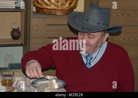 Testa e la spalla ritratto di sorridere uomo maturo in grigio il cappello da cowboy e maroon pullover è mangiare il dessert con una forchetta in home interni. Foto Stock