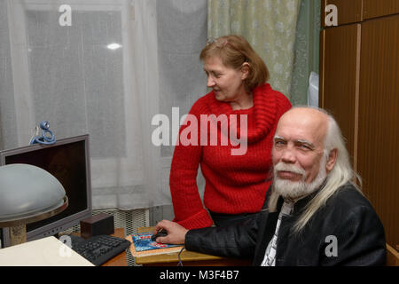 Donna matura in maglione rosso è in piedi vicino a sorridere senior uomo calvo con lunghi capelli grigi e la barba che è seduto in nero in corrispondenza del computer nella camicia Foto Stock
