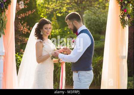 Groom anello di usura nel dito della sua sposa unter l'arco con pesca tessile di colore Foto Stock