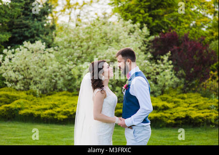 Bella lo sposo e la sposa nel parco tenendo le mani e guardando ogni altro. Appena sposato coppia giovane a baciare sulla natura verde dello sfondo. Foto Stock