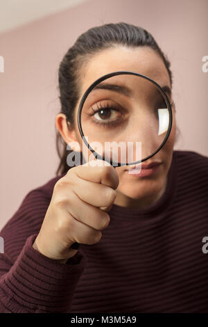 Donna alla ricerca di indizi o condurre un' indagine o ricerca il peering attraverso un palmare la lente di ingrandimento , ciò che rende il suo occhio sembrano più grande Foto Stock