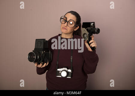 Da stupidi femmina appassionato di fotografia indossando grandi bicchieri con tre telecamere vintage con in mano una videocamera e fissando con spazio di copia Foto Stock