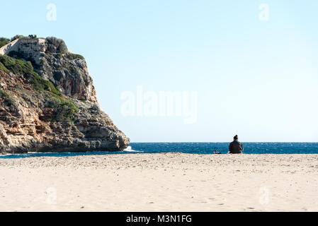 Silhouette di una donna seduta sulla sabbia della spiaggia e di fronte al mare a Cala Romantica sull'isola di Mallorca, Spagna. Foto Stock